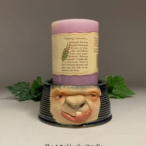 May include: A lavender-colored pillar candle sits in a unique ceramic holder. The holder features a sculpted face with a wide grin and a single tooth. The candle has a label that reads "Relaxing Lavender". The base of the holder is dark blue with ribbed detailing.