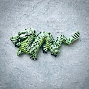 May include: A green ceramic dragon figurine with a detailed, scaled body, red eyes, and black claws. The dragon is posed in a curved position, with its head raised and tail curled. The figurine is on a textured, light blue surface.
