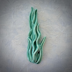May include: A vertical ceramic decorative piece in a glossy seafoam green glaze. The object features a series of elongated, undulating, flame-like or seaweed-like vertical ridges that create a sense of fluid motion and depth across the surface.