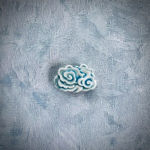 May include: A small, ceramic cloud-shaped decorative object. The cloud is primarily white with blue accents, featuring a swirling design. The object is set against a textured, light blue background.