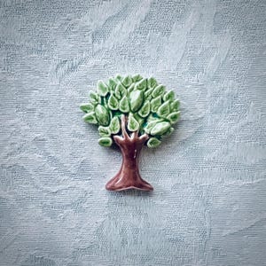 May include: A ceramic brooch shaped like a tree with a glossy brown trunk and a textured canopy of light and dark green leaves. The piece features a smooth, reflective finish on the surface of the leaves and the trunk, creating a dimensional look.