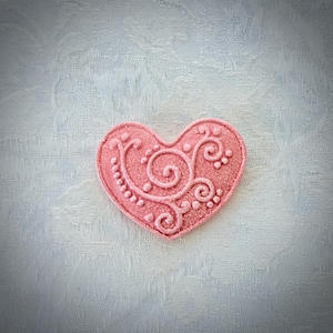 May include: A heart-shaped object in a soft pink hue, featuring a raised, intricate relief pattern of swirling lines and small dots across its surface. The texture appears slightly granular, and the design is symmetrical in its decorative filigree style.