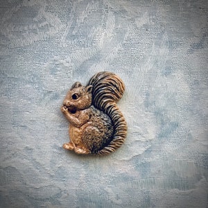 May include: A brown and white ceramic squirrel figurine with a bushy tail. The squirrel is sitting on a white textured surface.