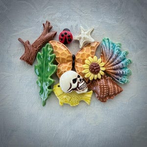 May include: A collection of small ceramic decorative pieces including a brown tree branch, a green leaf, a red ladybug, a white starfish, an orange butterfly, a yellow sunflower, a white skull, a yellow fish, and a brown seashell.