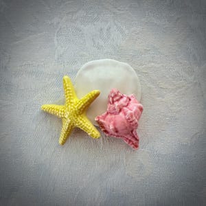 May include: A decorative arrangement consisting of a bright yellow five-pointed starfish, a smooth white circular sand dollar, and a glossy pink conch shell. These three distinct marine-themed items are clustered together in a single group.