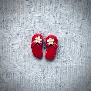 May include: Two red flip-flops with white flower accents. These miniature sandals are a charming accessory, perfect for summer-themed crafts or jewelry. The flip-flops are approximately 1 inch long.