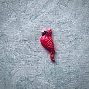 May include: A bright red cardinal bird brooch with a black mask around the eyes and a small orange beak. The bird is depicted in profile, facing left, with detailed molded feathers on its wings and a long, pointed tail extending downward.
