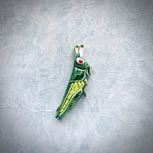 May include: A ceramic grasshopper figurine in shades of green and yellow, with red eyes and white antennae. The grasshopper is detailed with a glossy finish, and is set against a textured, light-colored background.