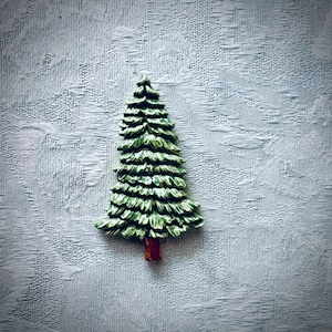 May include: A ceramic Christmas tree figurine with a green and white glaze. The tree has a brown trunk and is set against a textured, light blue background. The figurine is a festive holiday decoration.