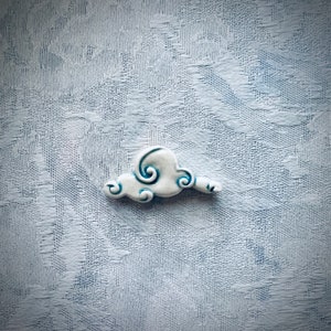 May include: A small, white ceramic cloud with blue accents. The cloud has a whimsical design with swirling details and a light blue outline. The background is a textured, light blue fabric.