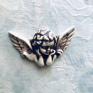 May include: A white ceramic angel figurine with brown painted details. The angel is depicted with wings and is looking down with its head resting on its hand.