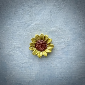 May include: A ceramic sunflower with yellow petals and a brown center. The flower is set against a light blue textured background. The sunflower is a decorative item.