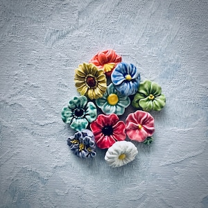 May include: A collection of colorful ceramic flower-shaped buttons. The buttons are in various shades of blue, green, yellow, red, and purple.