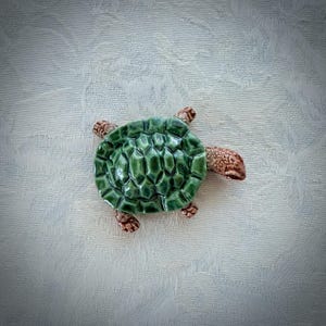 May include: A ceramic turtle figurine featuring a glossy, deep green shell with a textured, raised geometric pattern. The head, legs, and tail are finished in a mottled, light brown glaze with a speckled appearance, showing distinct sculpted details.