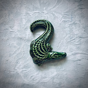 May include: A green ceramic alligator figurine with a textured, scaly design. The alligator is curled into a 'C' shape with its tail curled up and its head facing the right.