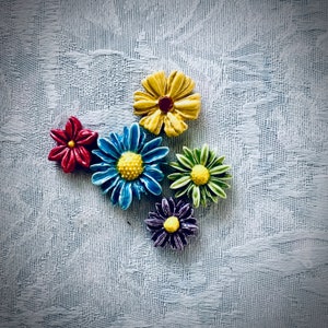 May include: A collection of six ceramic flower-shaped decorative items in various colors. The flowers include red, blue, yellow, green, and purple, each with a contrasting center. The items are arranged on a textured, light blue surface.