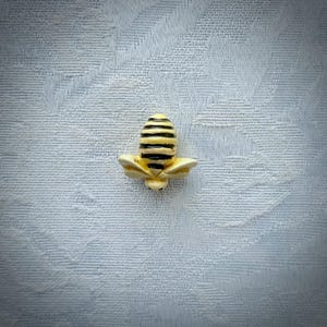 May include: A small, detailed bee-shaped pin. The bee has a yellow body with black stripes and delicate, cream-colored wings. The pin is likely made of metal or ceramic and is set against a light blue textured background.