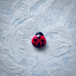 May include: A red and black ladybug figurine with black spots. The ladybug is sitting on a white textured surface.