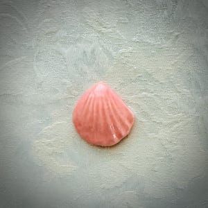 May include: A small, pink ceramic seashell with a glossy finish. The shell has a ridged texture and is set against a light-colored, patterned background. The seashell is a decorative item.