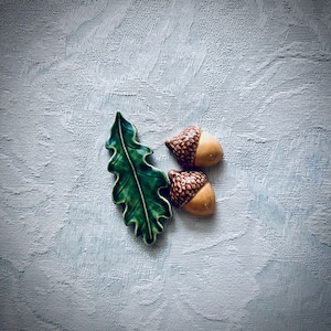 May include: A ceramic green oak leaf figurine positioned next to two small ceramic acorns. The leaf has a glossy finish with defined veins, while the acorns feature textured brown caps and smooth, light tan bases, creating a nature-inspired set.