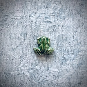 May include: A green ceramic frog figurine with a textured surface. The frog is sitting on a white textured background.
