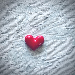 May include: A glossy, red heart-shaped object rests on a textured, light blue surface. The heart is smooth and reflects light, creating highlights. The background has a subtle, patterned design, adding depth to the image. The heart is a symbol of love.