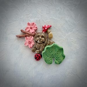May include: A ceramic brooch featuring a brown dog sitting next to a red and white spotted mushroom, a large green leaf, a small red ladybug with black spots, and a brown branch with two pink flowers with five petals each.