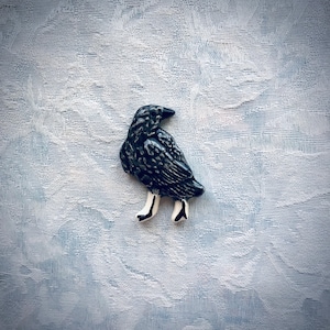May include: A ceramic raven figurine with a detailed, textured black body and white legs with black accents. The bird is perched on a light blue and white textured surface. The raven is a decorative item.