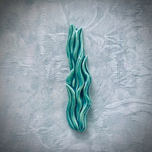 May include: A teal ceramic sculpture with a flowing, organic form. The glossy finish highlights the curves and ridges of the piece. The sculpture is a decorative item, possibly for a home or office setting.