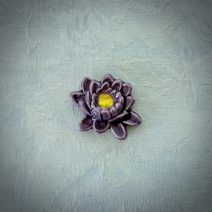 May include: A ceramic flower ornament shaped like a water lily with multiple layered petals in a glossy dark purple finish. The center of the flower features a bright yellow circular accent, providing a distinct contrast to the surrounding purple petals.