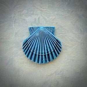 May include: A bright blue ceramic scallop shell ornament featuring a glossy finish and distinct, raised radial ridges that extend from the hinge to the scalloped outer edge, creating a symmetrical fan-shaped design with deep grooves.