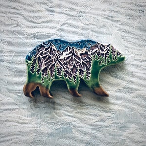 May include: A ceramic bear-shaped pin featuring a detailed relief carving of a mountain range and pine forest. The design incorporates a blue sky at the top, white snow-capped peaks, green trees, and a brown base, all finished with a glossy glaze.