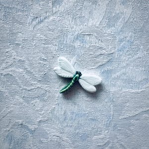 May include: A white dragonfly figurine with detailed wings and a green body rests on a textured, light blue surface. The dragonfly's wings are delicately sculpted, and the body has a glossy finish. The figurine is a decorative item.