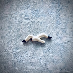 May include: A white ceramic seagull figurine with black painted wings and beak, flying in a diving pose. The figurine is on a white textured background.