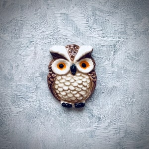 May include: A detailed owl figurine with a brown, white, and cream color scheme. The owl has large orange eyes and a black beak. The figurine is set against a textured, light blue background.