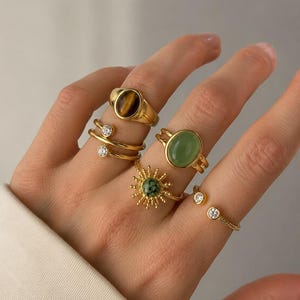 Puede incluir: Una colección de cinco anillos dorados en los dedos. El conjunto incluye un anillo con una piedra ojo de tigre ovalada, un anillo con una piedra verde ovalada, un anillo con forma de sol y una piedra verde, y dos anillos abiertos con pequeñas piedras claras.