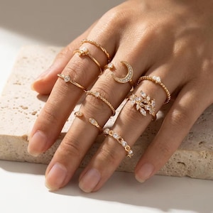 Puede incluir: Un conjunto de anillos dorados en los dedos con varios diseños, incluyendo una media luna, un motivo de hojas con piedras transparentes, pequeñas perlas, bandas trenzadas y anillos delicados con piedras brillantes individuales.