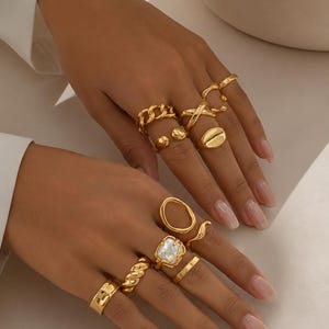 Puede incluir: Una colección de diez anillos dorados usados en los dedos, con varios diseños que incluyen una banda de eslabones de cadena, un anillo de sello ovalado, un anillo cuadrado con incrustación de piedra blanca, una banda trenzada y varios anillos lisos, abiertos y cruzados.
