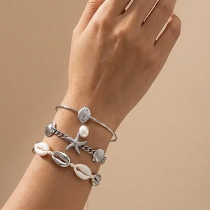 Puede incluir: Un conjunto de tres pulseras con elementos de metal plateado que incluyen una estrella de mar, dijes de conchas y una perla blanca. Una pulsera es un brazalete rígido, otra es de cadena y la tercera es un cordón con conchas.
