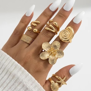 Puede incluir: Una colección de anillos dorados con diversos diseños, incluyendo una flor grande de cinco pétalos, una concha en espiral, una estrella de mar, una concha más pequeña, una banda texturizada y dos anillos abiertos con extremos redondeados.