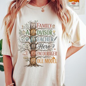 Op de afbeelding: Een crèmekleurig t-shirt met een afbeelding van een boom waarvan de stam bestaat uit gestapelde blokken die FATHER spellen. De takken tonen de woorden FAMILY, ADVISOR, TEACHER, HERO, ENCOURAGER en ROLE MODEL, vergezeld door kleine iconen.
