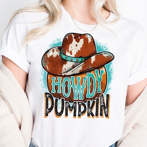 Peut inclure: Un t-shirt blanc avec un motif représentant une citrouille orange portant un chapeau de cowboy à taches de vache marron et blanc. Le texte HOWDY PUMPKIN est imprimé en lettres turquoise et noires sur la citrouille, sur un fond circulaire bleu sarcelle.