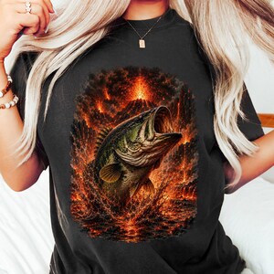 May include: A black short-sleeve t-shirt featuring a graphic print of a large green bass fish leaping out of water. The background of the graphic depicts a volcanic eruption with bright orange and red lava flowing down dark rocky mountains.