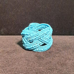 May include: A multi-strand bracelet constructed from numerous small, round, bright turquoise blue beads. The beads are arranged in a woven, crisscross pattern, creating a thick, textured, and circular band structure with a uniform colour throughout.