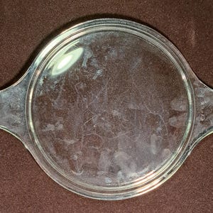 Puede incluir: Una tapa de cristal Pyrex circular y transparente con dos asas laterales integradas. La superficie del cristal es transparente con reflejos de luz visibles y manchas tenues. La palabra Pyrex está grabada en una de las asas laterales.