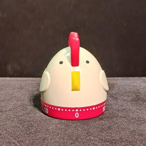May include: A kitchen timer shaped like a chicken with a cream-colored body, a bright red comb, and a yellow beak. The base is a red rotating dial featuring white numbers and markings, with the number 0 centered at the front of the device.