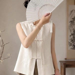 May include: A sleeveless off-white vest featuring a subtle tonal floral pattern, a high collar, and front closures with decorative pearl-like buttons and long hanging cords. The vest is paired with a white folding hand fan with a tassel.