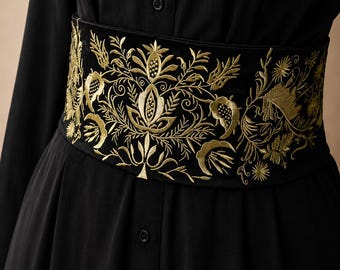 May include: A wide black belt featuring intricate gold embroidery in a floral and botanical pattern. The belt is worn over a black button-down dress. The embroidery consists of stylized leaves, stems, and flower motifs that wrap around the waist.