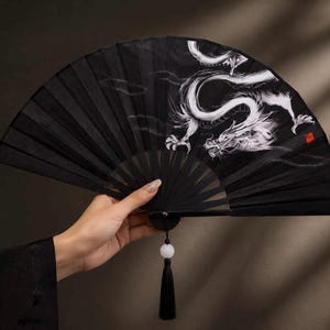 May include: A black folding hand fan featuring a white dragon illustration with flowing lines. The fan includes a black handle, a white bead, and a black tassel hanging from the base. A small red square seal is visible on the right side.