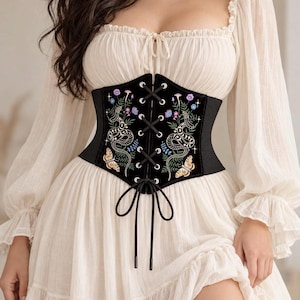 May include: A black corset belt featuring intricate embroidery of snakes, moths, and wildflowers. The front panel has a lace-up design with silver-toned metal grommets and black cord ties. The side panels are made of a ribbed elastic material.